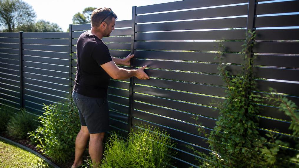 A homeowner installs a slatted aluminium fence in a sunny, green backyard.